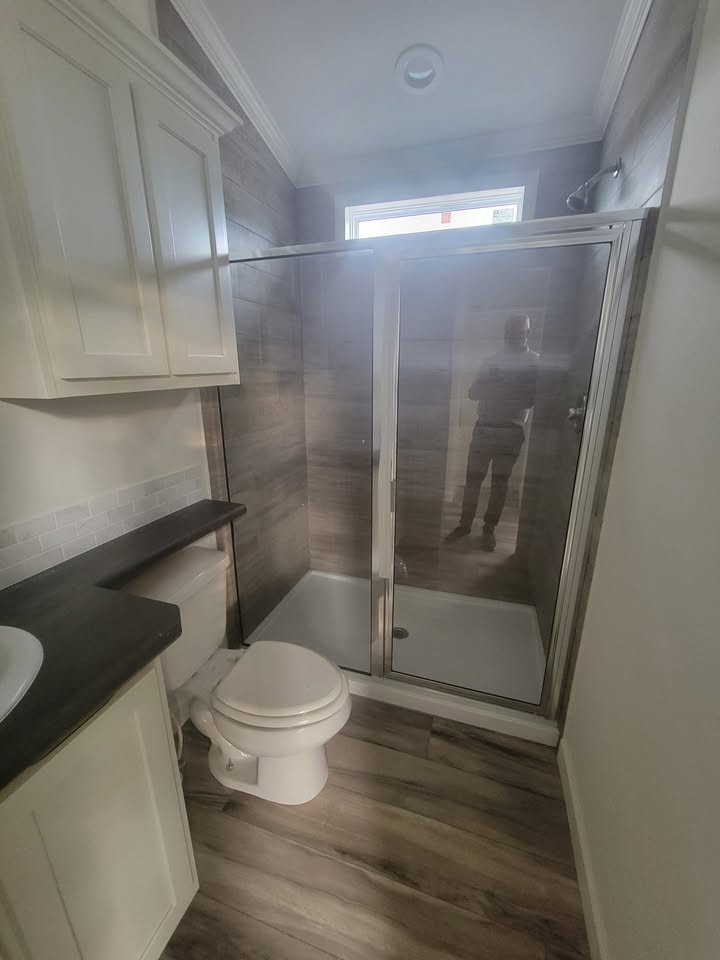 A small bathroom features a glass-enclosed shower, a white toilet, and a dark countertop with a sink. A cabinet is mounted above, and light wood flooring adds warmth.