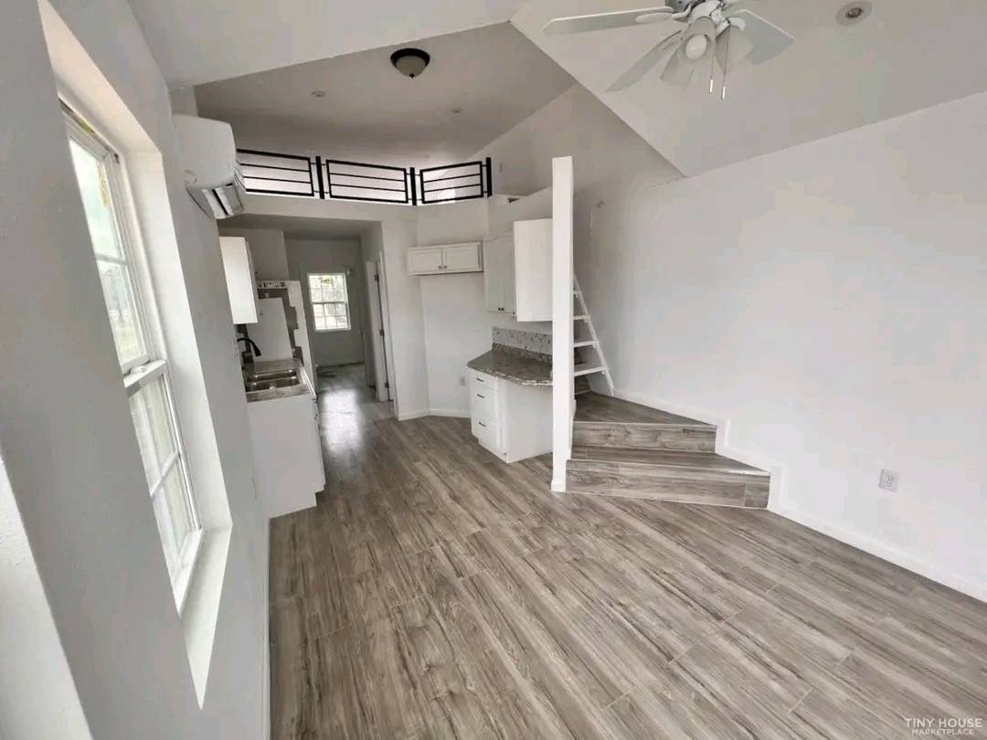 Cozy modern tiny house interior with white walls and wood flooring. Features a compact kitchen, spiral staircase, loft area, and ceiling fan. Bright and airy.