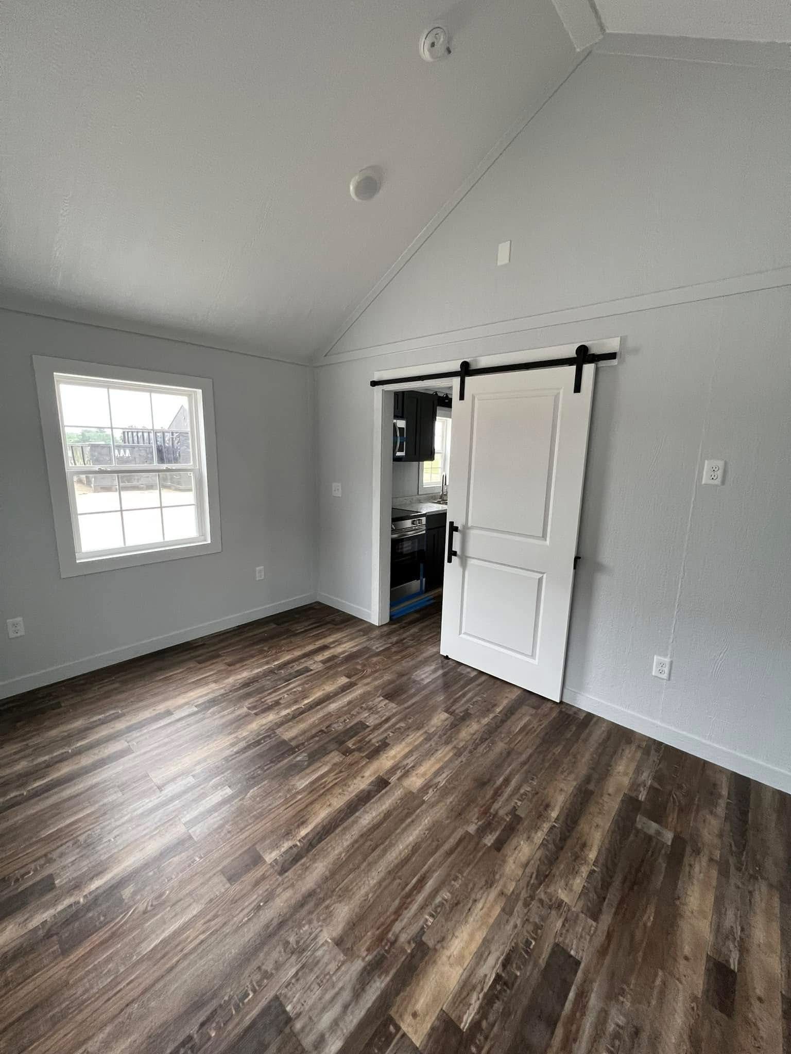A small, empty room with light gray walls, a wood floor, and a window on the left. A white sliding barn door on the right reveals a kitchen area.