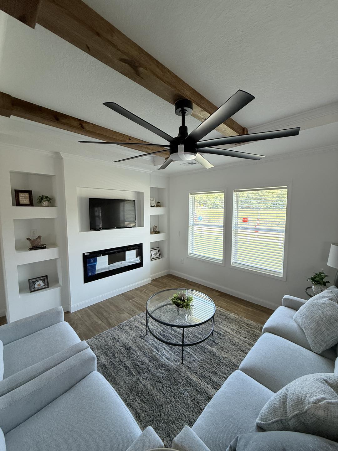 Modern living room with white walls and a wooden-beam ceiling. It features a large fan, TV, fireplace, round glass table, two sofas, and large windows. Cozy ambiance.