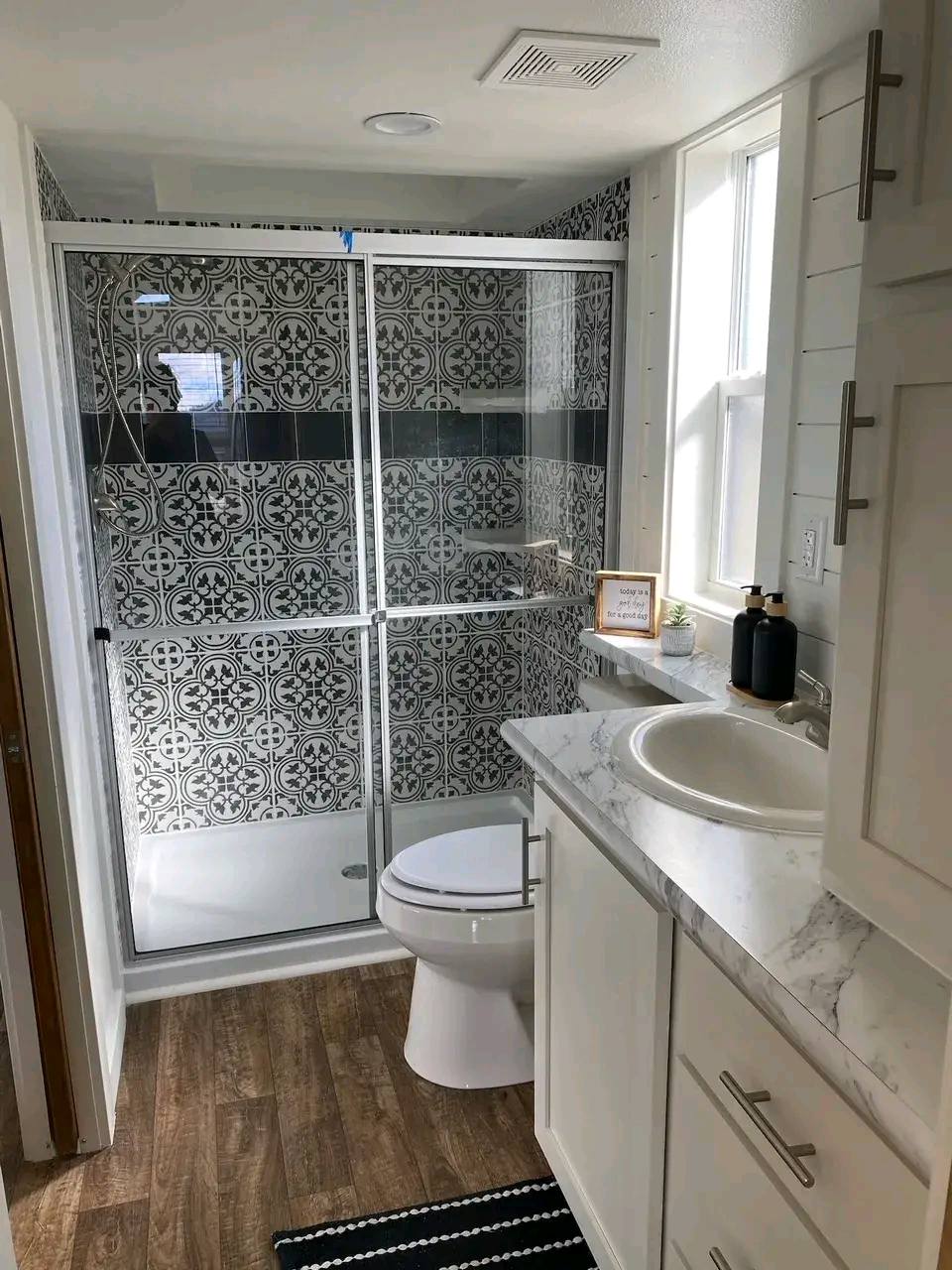 Stylish bathroom with patterned black and white tile shower, glass door, wood floor, white vanity with marble top, and decorative accents. Bright, modern feel.