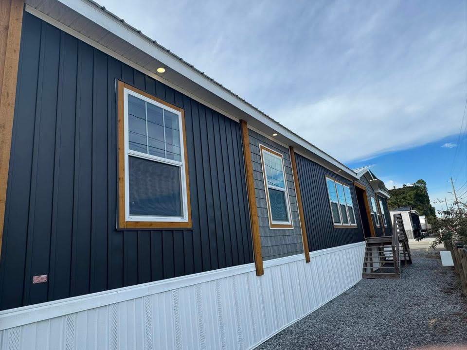 A modern, dark-paneled modular home with wooden trim and white lower siding. The sky is partly cloudy, and a gravel path leads to the entrance.