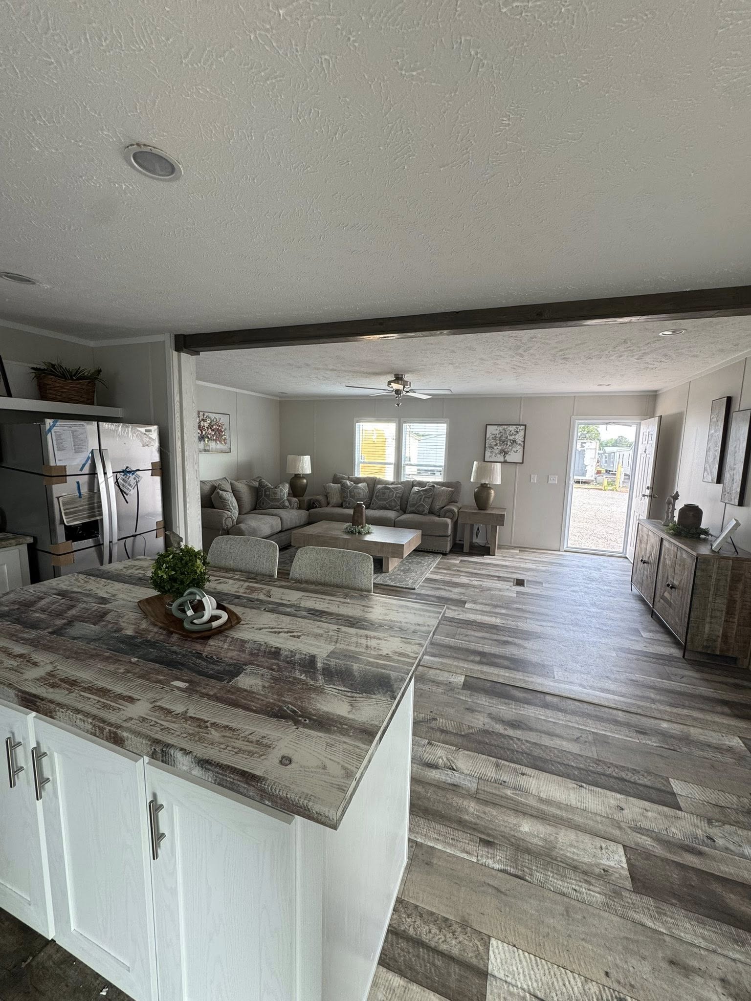 Open living room with rustic wooden flooring and a white kitchen island. Features a large sectional sofa and paintings on light gray walls, conveying a cozy, modern ambiance.