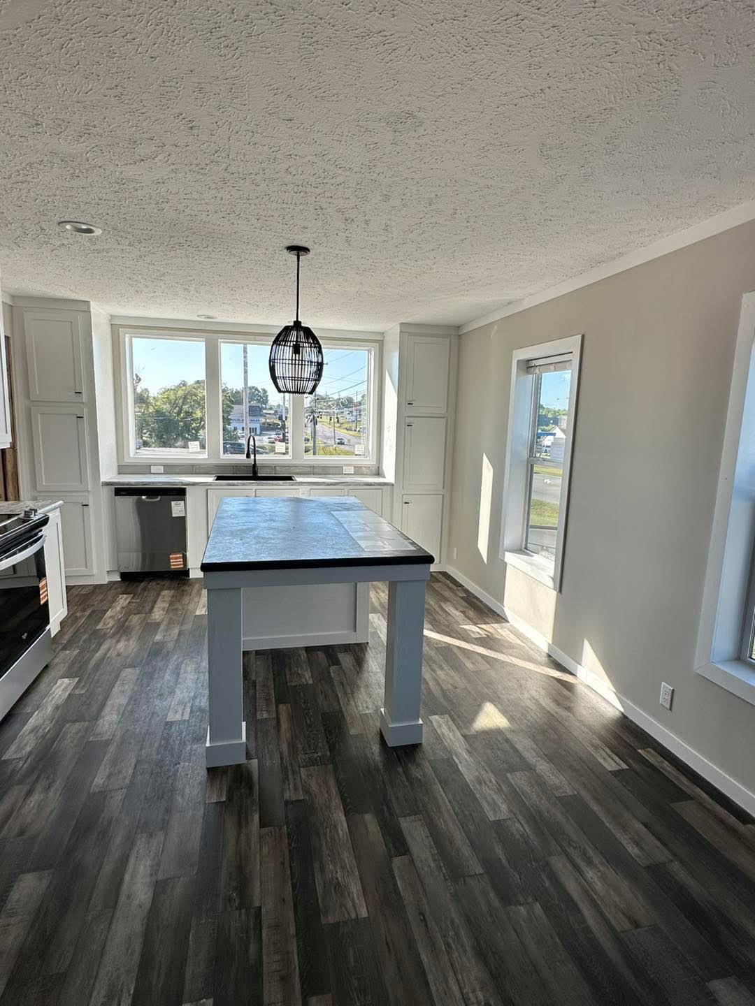 Bright kitchen with large windows, white cabinets, and dark wood flooring. Features a central island with a modern pendant light, creating an airy, inviting space.