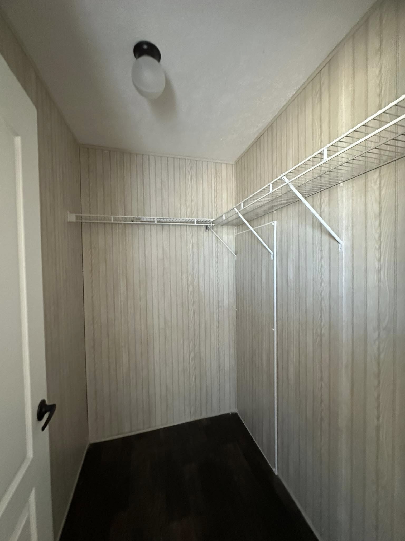 Empty walk-in closet with light wood panel walls, dark flooring, and white wire shelving on two sides. A simple ceiling light provides illumination.