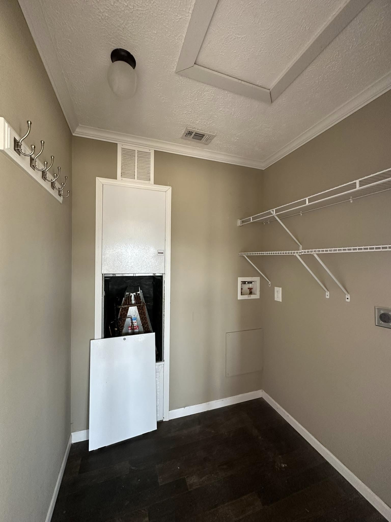 Narrow utility room with beige walls and dark wood floor. White wire shelves line the right wall. A panel is detached on the far wall.