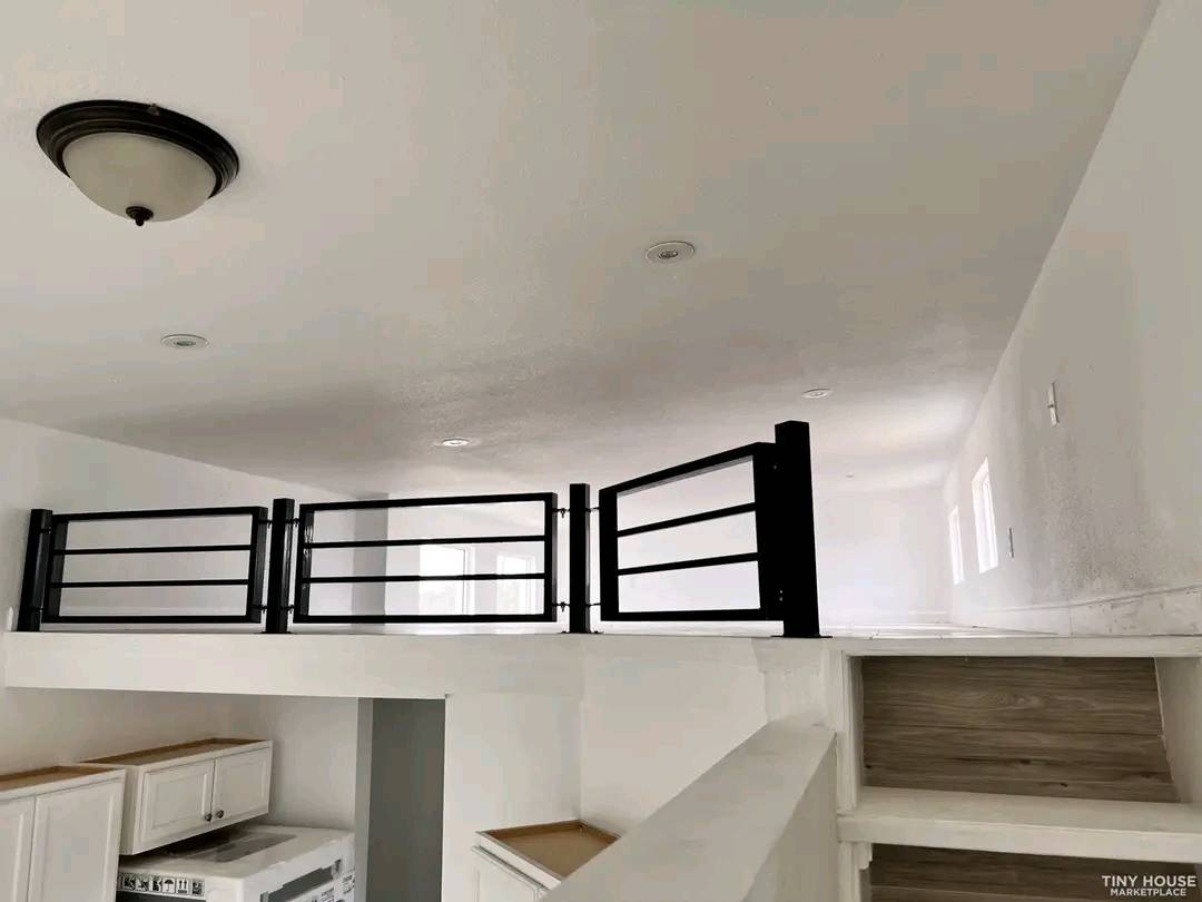 A minimalist white tiny house interior featuring a loft with a black railing. Stairs lead up, and kitchen cabinets are partially visible below. Bright and airy.
