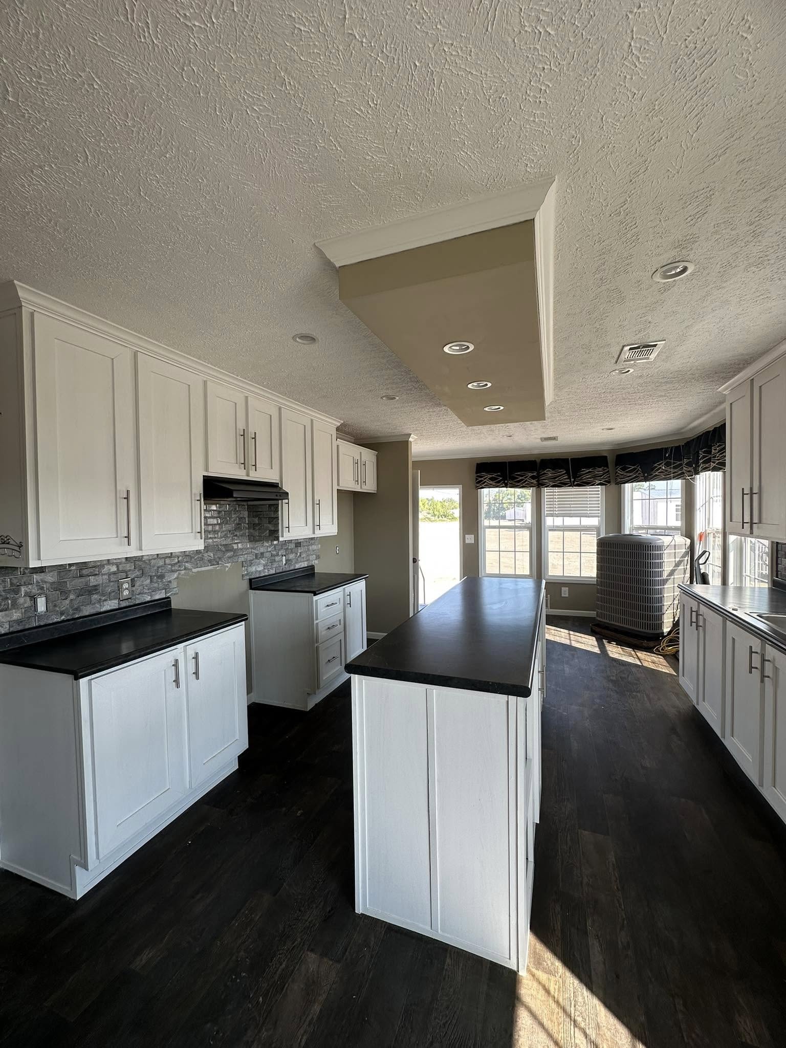 Modern kitchen with white cabinets, black countertops, and dark wood floors. A large island centers the room with natural light from wide windows.