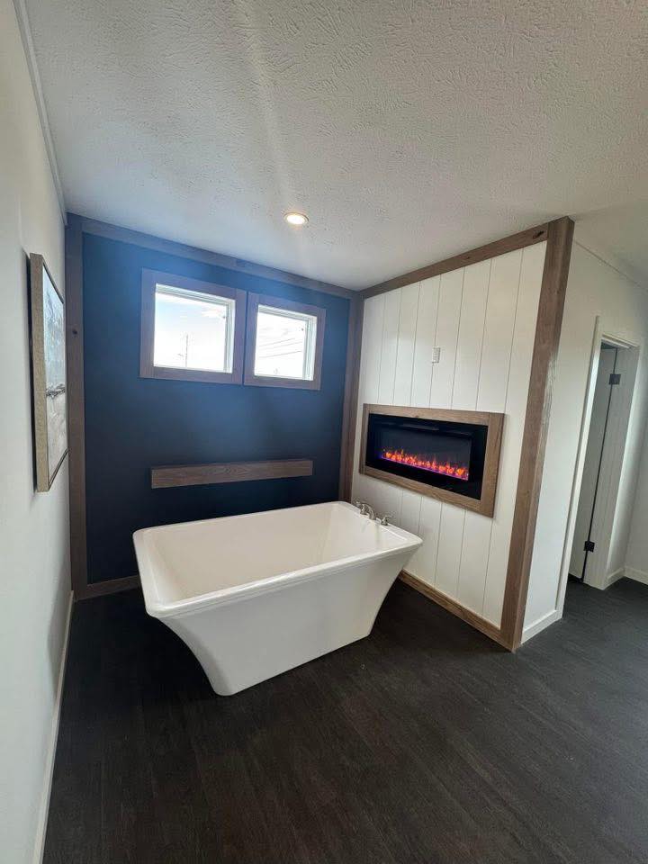 A modern bathroom features a freestanding white tub, dark wood flooring, and a sleek electric fireplace on a white-paneled wall, evoking warmth and relaxation.