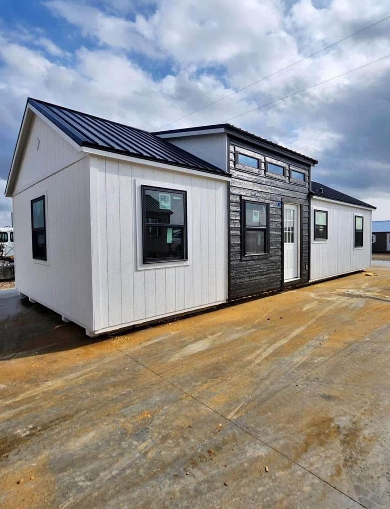 Modern tiny house with a black metal roof and a mix of white and dark wood siding against a partly cloudy sky. Windows and a white door are visible.