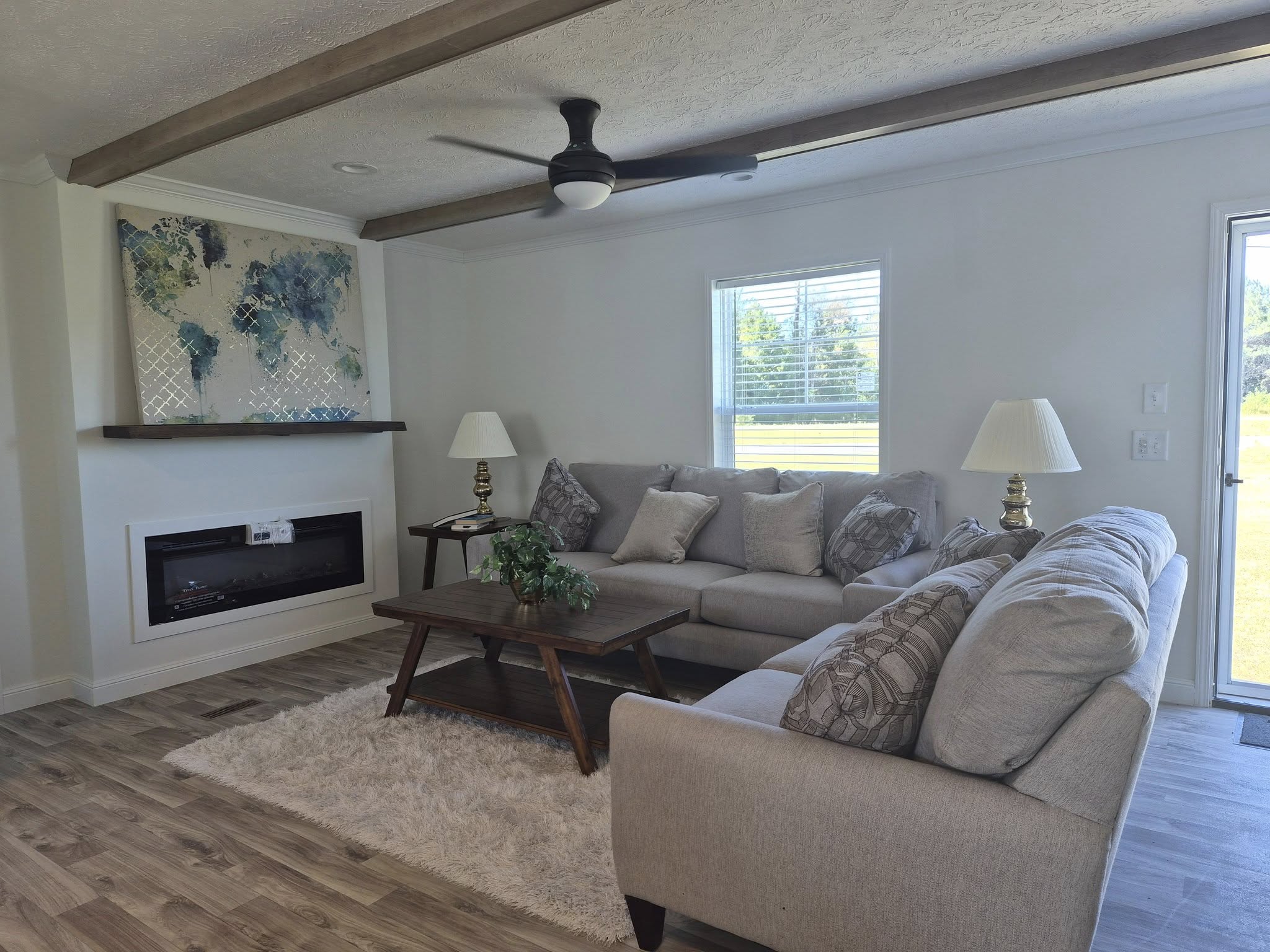 Modern living room with gray sectional sofa, wooden coffee table, and abstract artwork above a sleek fireplace. Ceiling fan and large window add light.