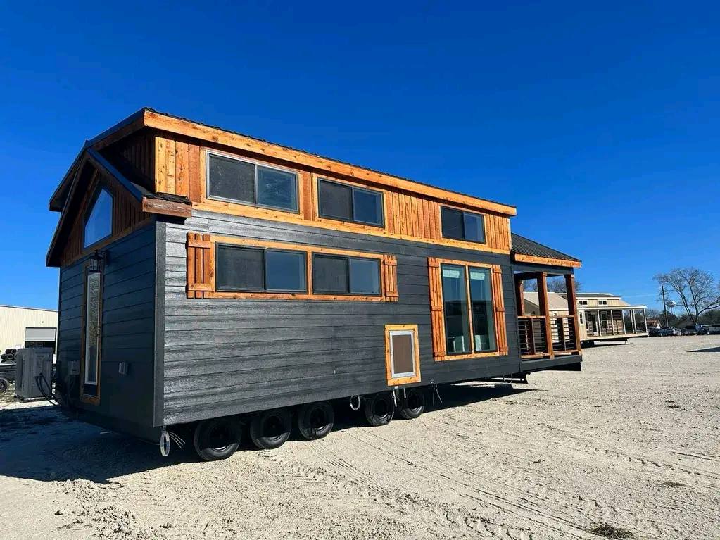 A tiny house on wheels with a rustic design, featuring wood accents and large windows. It sits on a sunny, open lot under a clear blue sky.