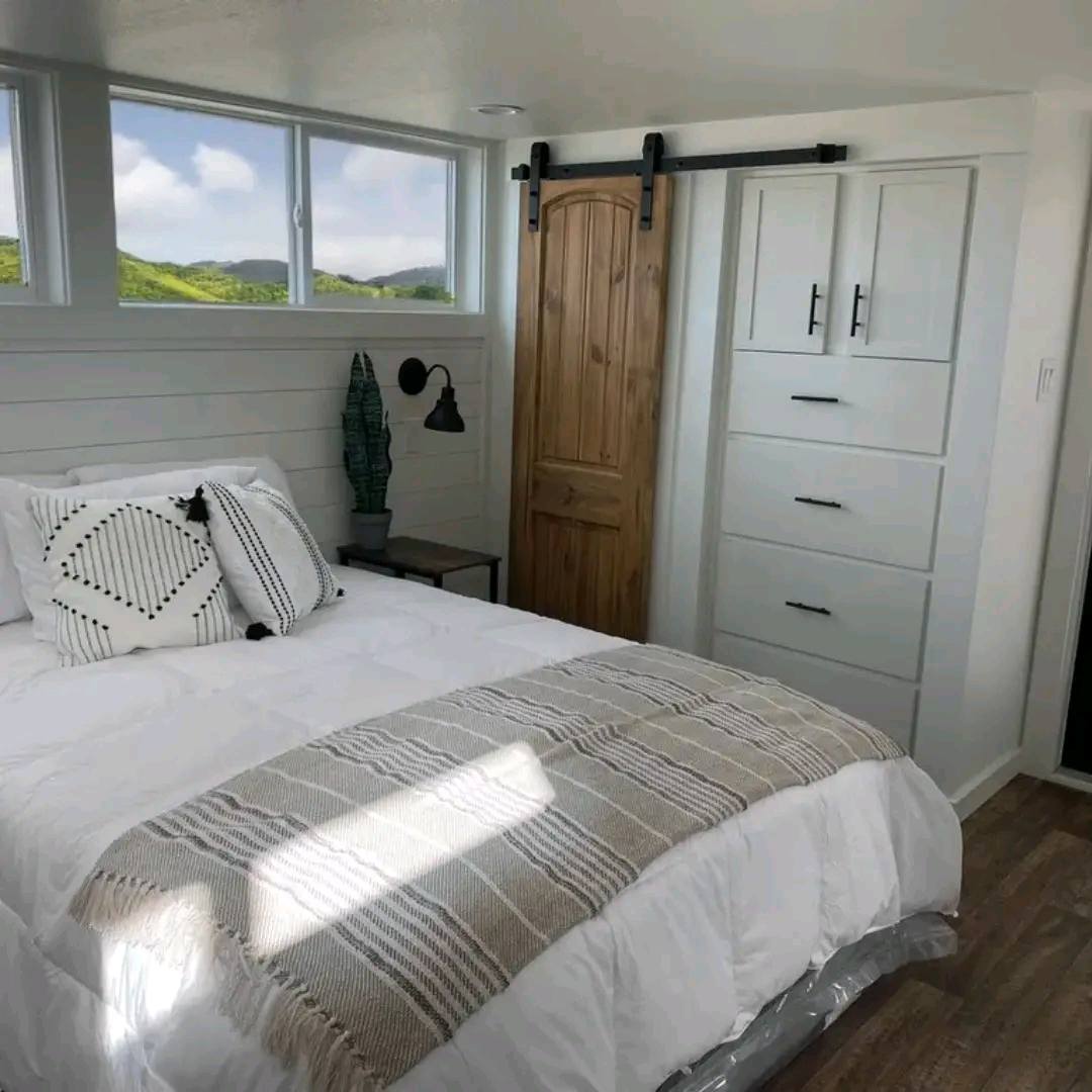Cozy bedroom with white shiplap walls, a large bed with striped pillows, a knit throw blanket, wooden barn door, dresser, and scenic window view.
