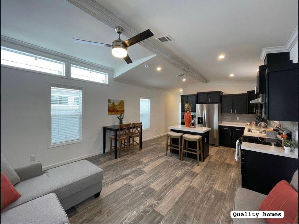 Modern open-plan kitchen and living room with gray furniture, wooden accents, and a ceiling fan. Stainless steel appliances enhance the sleek design.