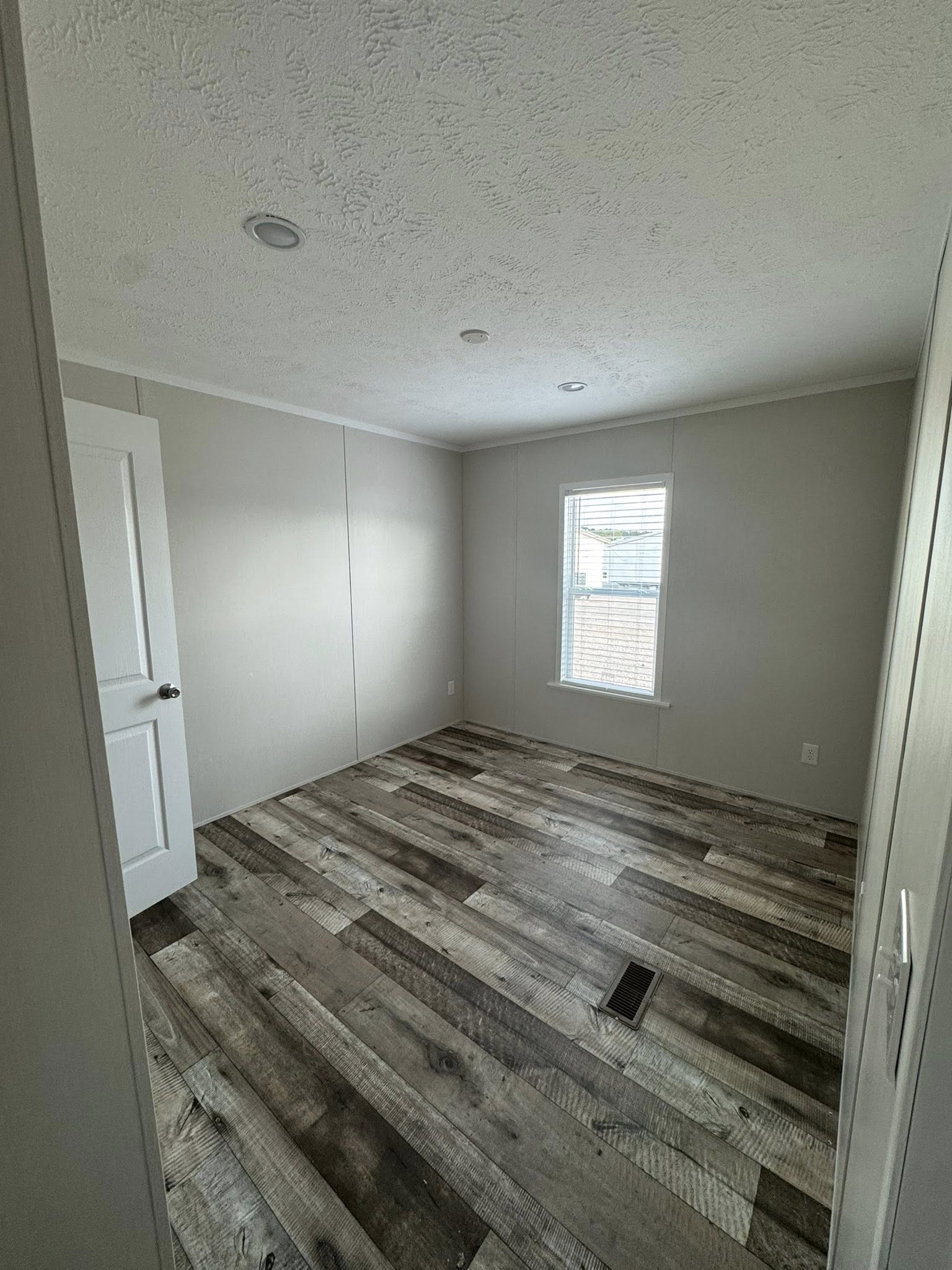Empty room with light gray walls and rustic, wood-textured flooring. A single narrow window lets in natural light. The atmosphere is minimalistic and serene.