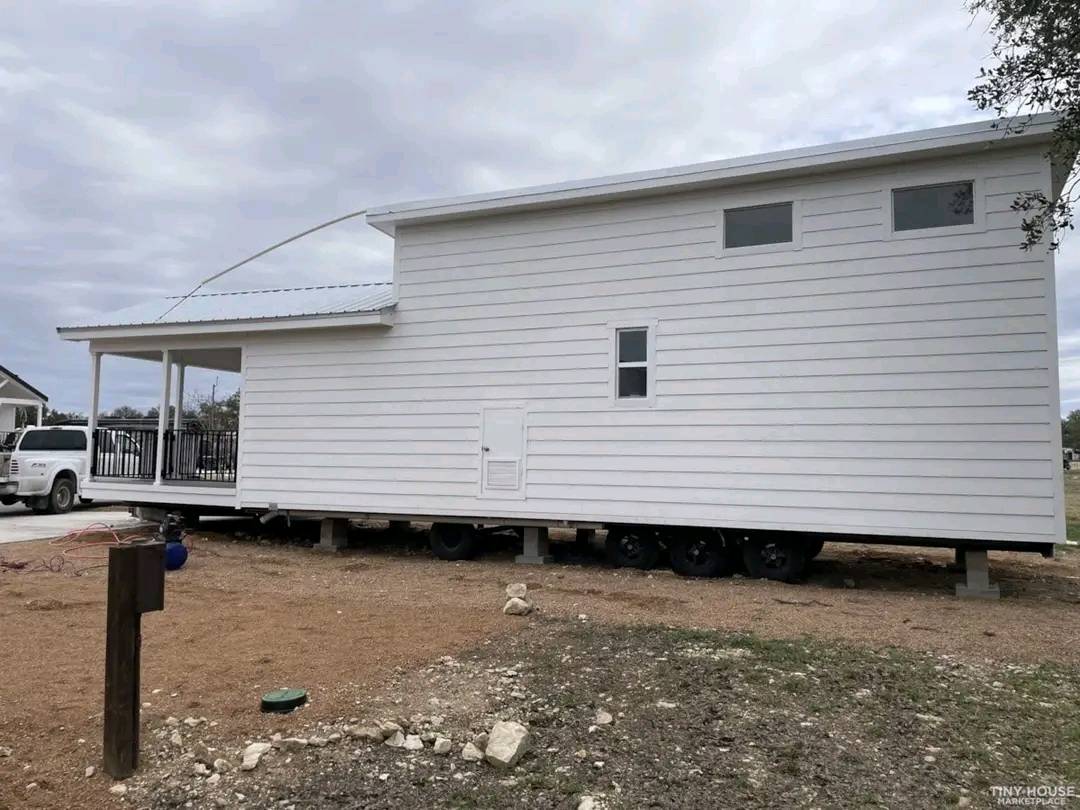A modern white tiny house on wheels is set on a gravel lot. It has a slanted roof, small windows, and a porch. A white vehicle is parked nearby.