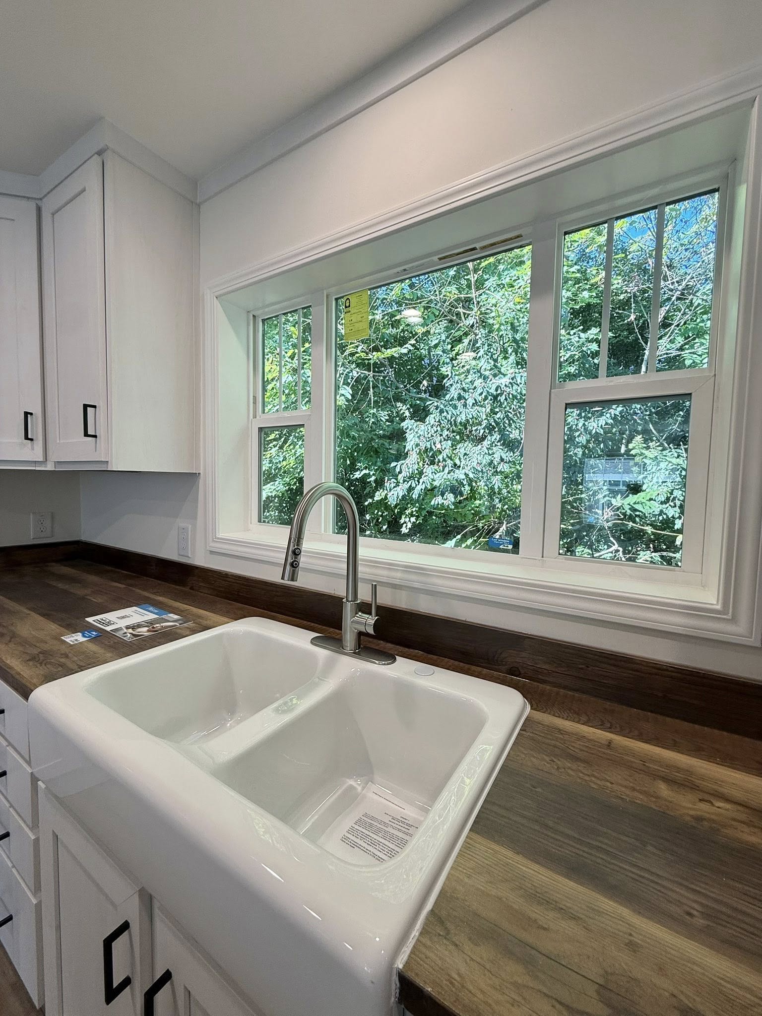 A bright kitchen with a large window overlooking lush greenery. A white double sink with a chrome faucet sits in a wooden countertop, creating a serene vibe.