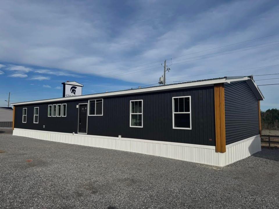 A long, dark gray modular building with white trim sits on a gravel lot under a partly cloudy sky. It has several windows and a small Spartan logo sign.