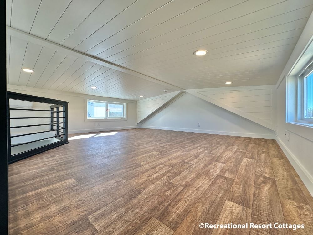 Spacious, serene attic room with wooden floors, sloped white ceiling, and recessed lighting. Two large windows add natural light. Airy and calm ambiance.