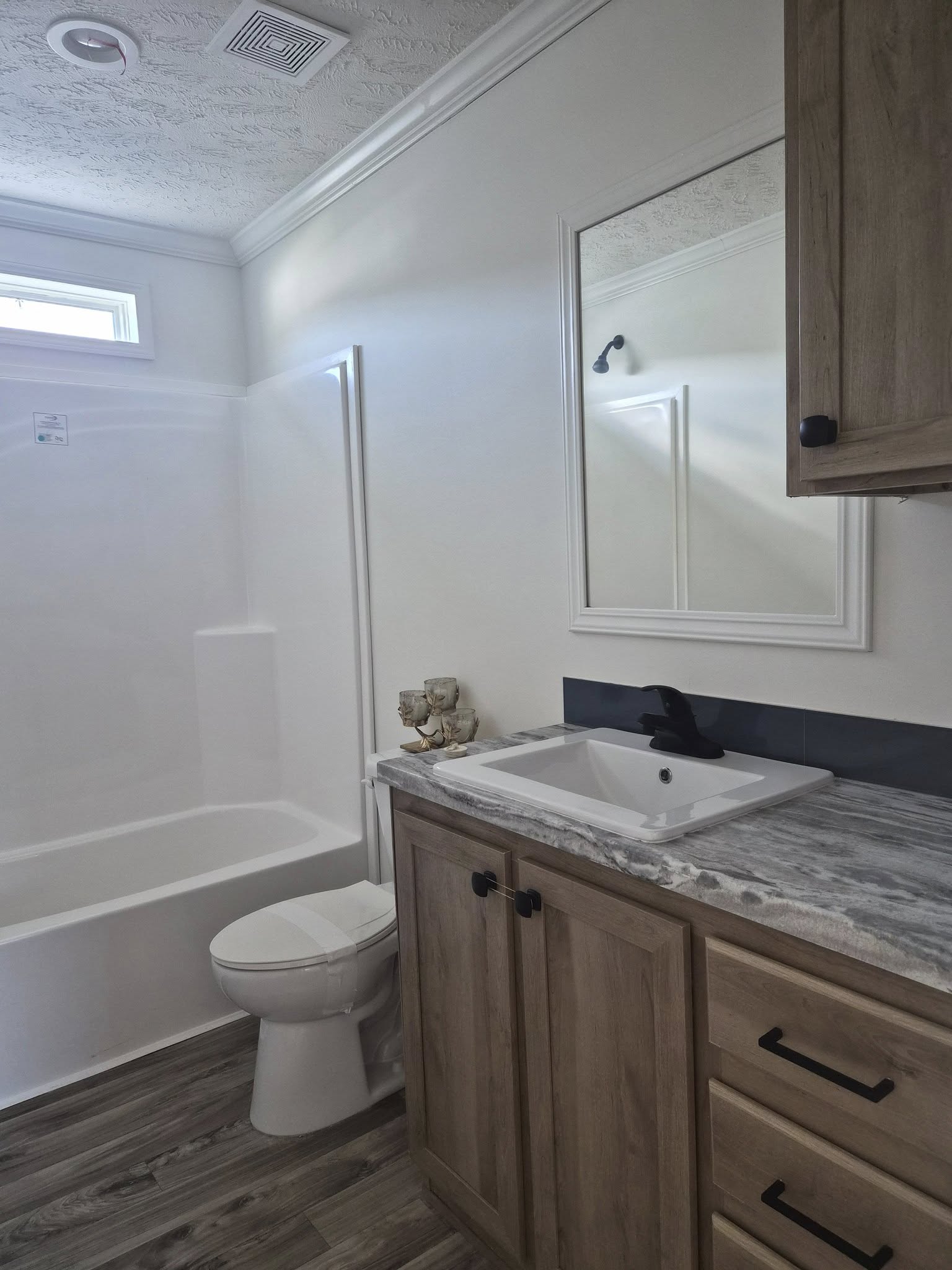 A compact bathroom with a shower-tub combo, white walls, and a small window. A wooden vanity with a marble countertop and black fixtures is on the right.