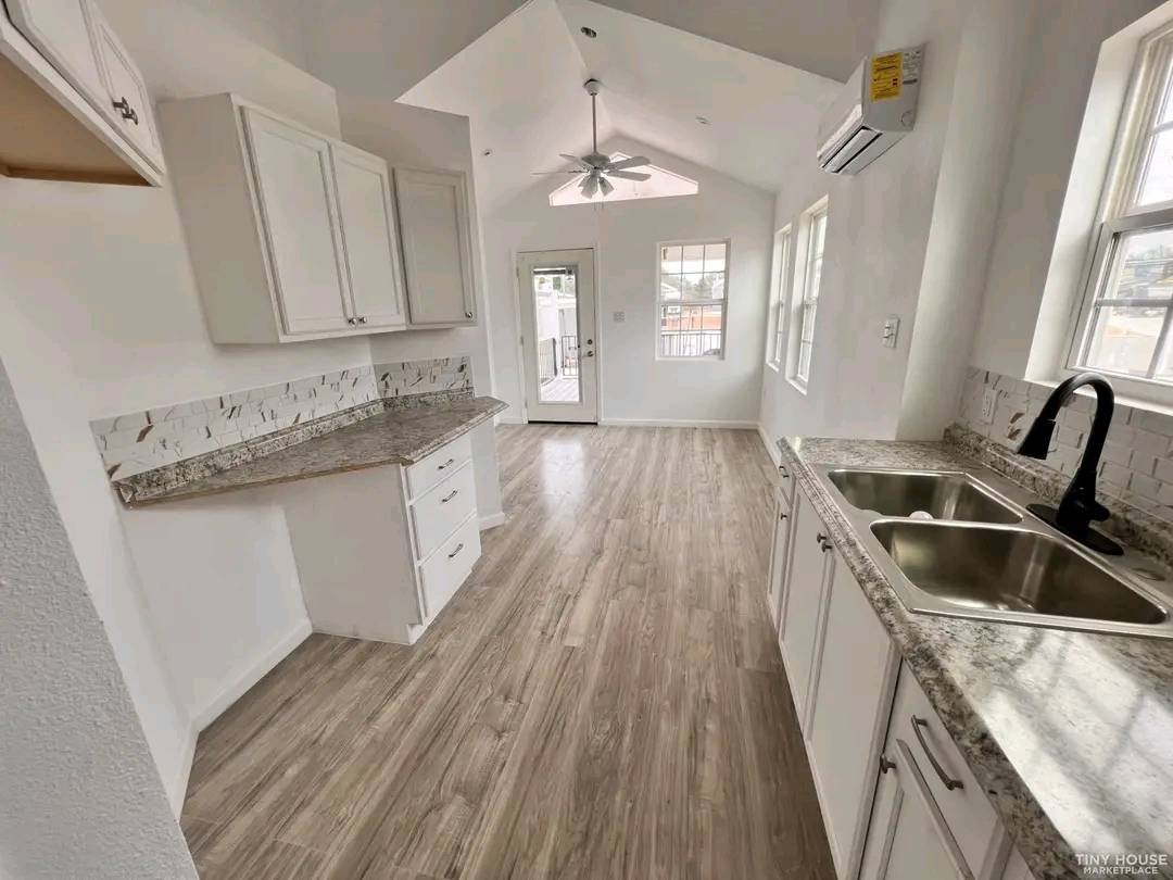 Bright, modern kitchen with light wood flooring, white cabinets, and granite countertops. Dual sink, ceiling fan, and windowed door leading outside. Airy and inviting tone.