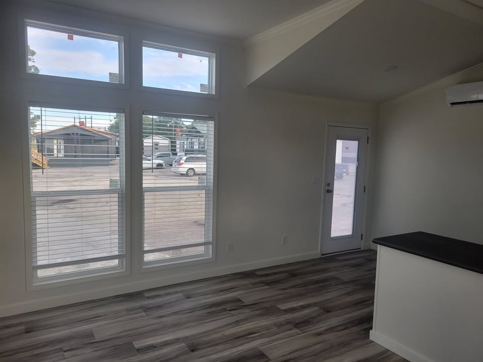 Bright room with large windows showing a view of a parking lot. Light wood flooring and a white wall with a small black counter. Airy and modern feel.