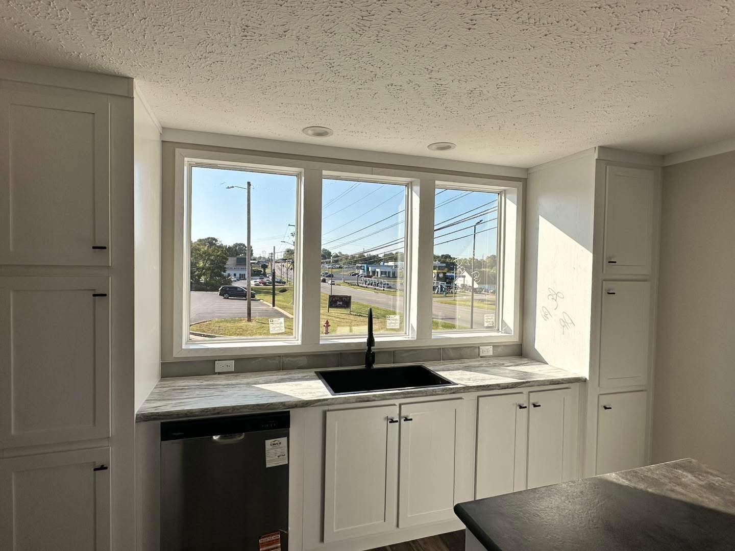 Bright kitchen featuring large windows with a view of the street. White cabinets, a black faucet, and a modern design create a fresh, inviting atmosphere.