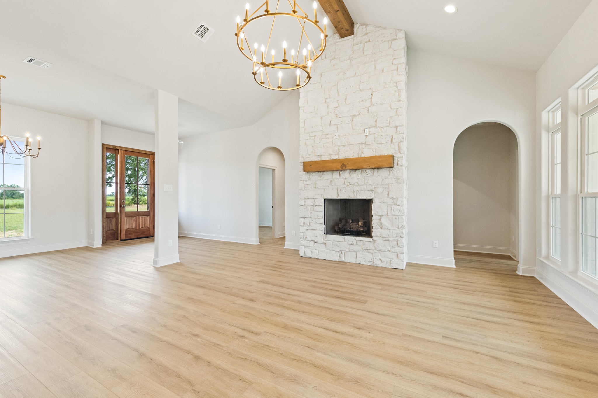 Spacious living room with light wood flooring and large windows. Features a white stone fireplace and two elegant chandeliers. Bright and airy ambiance.