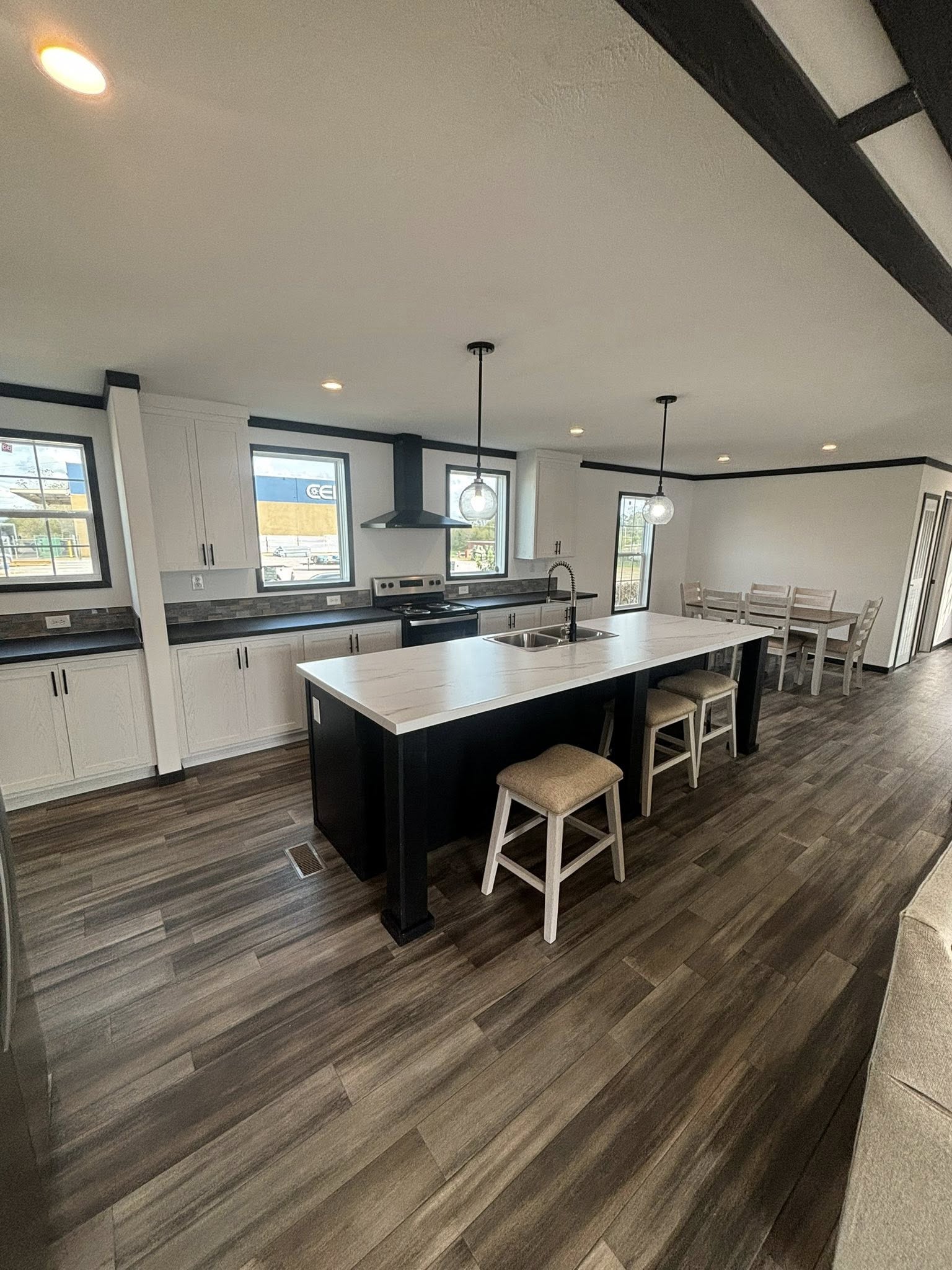 Modern kitchen with wood flooring, featuring a large island with a marble countertop, two stools, pendant lights, and a dining area in the background.