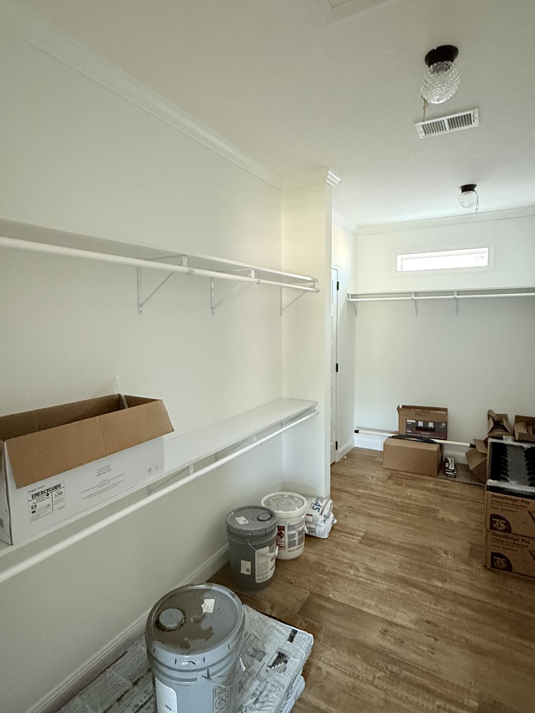 A spacious, empty closet with white shelves and hanging rods. Boxes, paint cans, and materials are scattered on the light wooden floor, suggesting a renovation in progress.