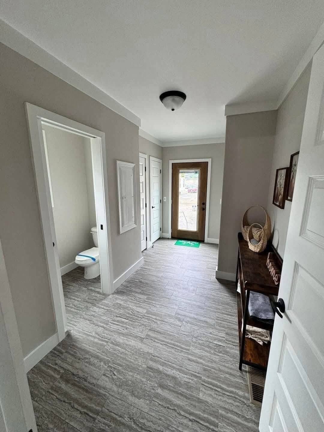 A hallway with light gray walls, a tiled floor, and white trim features a wooden door at the end. A bathroom is on the left, and a table with baskets and decor is on the right.