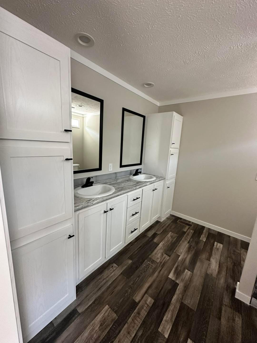 Modern bathroom with a double vanity featuring white cabinets and black hardware. Two rectangular mirrors hang above marble countertops. Wood flooring adds warmth.