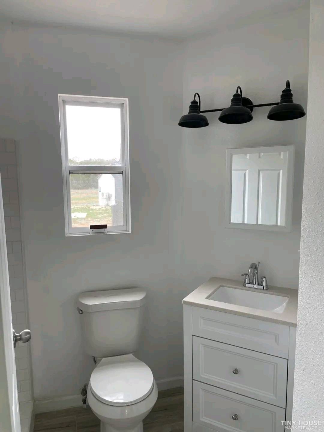 Compact bathroom with white walls and a small window, featuring a toilet and a white vanity with modern faucet. Three black lights above a mirror. Cozy and minimalist.