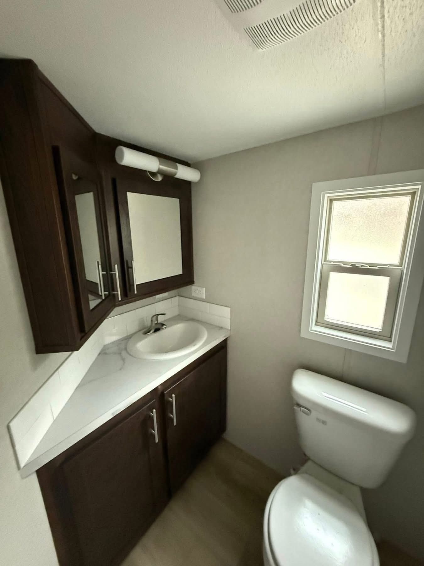 Compact bathroom featuring dark wood cabinetry with mirrored doors, a white sink, and a small window. The atmosphere is clean and minimalist.