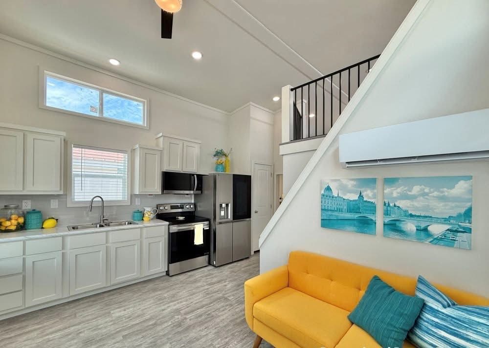 A modern kitchen with white cabinets and stainless steel appliances on the left, and a vibrant yellow couch with teal pillows on the right. Bright, spacious, and inviting.