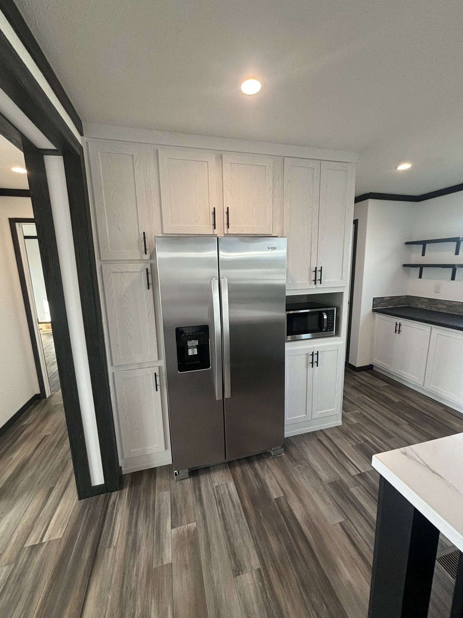 Modern kitchen with a stainless steel refrigerator, white cabinets, and a microwave. Features wood-patterned flooring and open shelving, creating a sleek and organized space.