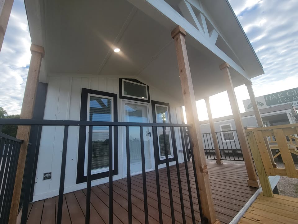 A modern porch with wooden floor and columns, black railing, and white siding, under a cloudy sky. Mood is inviting and serene.