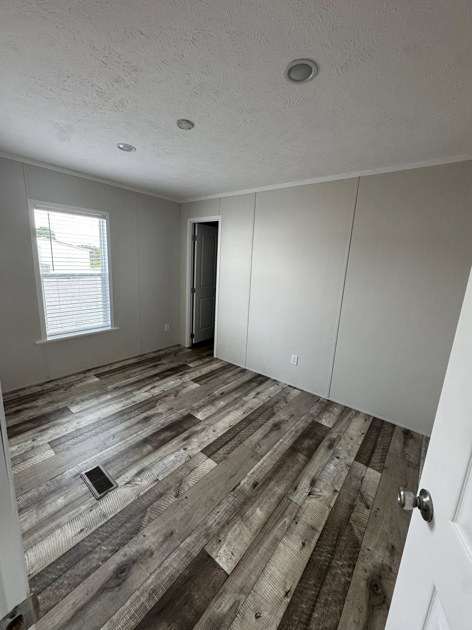 Empty room with light gray walls and rustic wood flooring. A closed door on the right, a window on the left with blinds, and recessed ceiling lights.