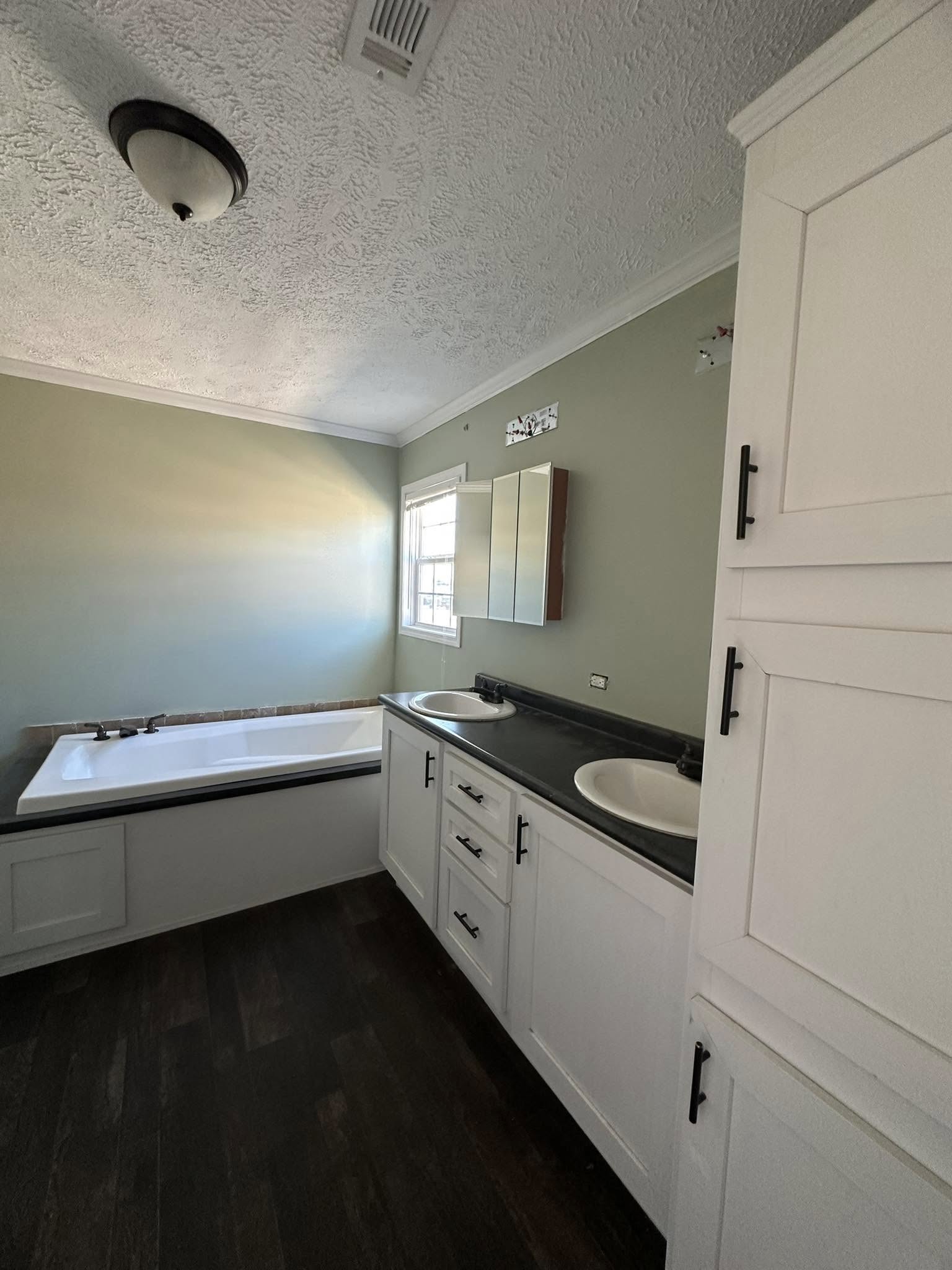 A modern bathroom with a white double vanity and black countertop, two sinks, a large bathtub, and green walls. Sunlight streams through a small window.