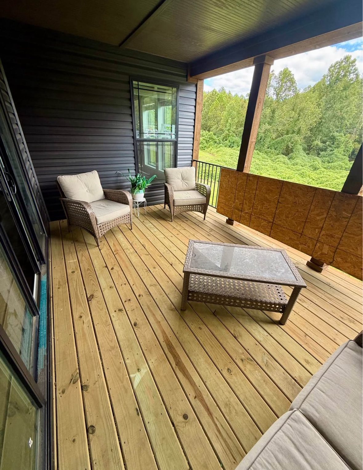 A cozy porch with wooden flooring features two wicker chairs with cushions, a glass-top table, and lush green trees in the background, creating a serene setting.