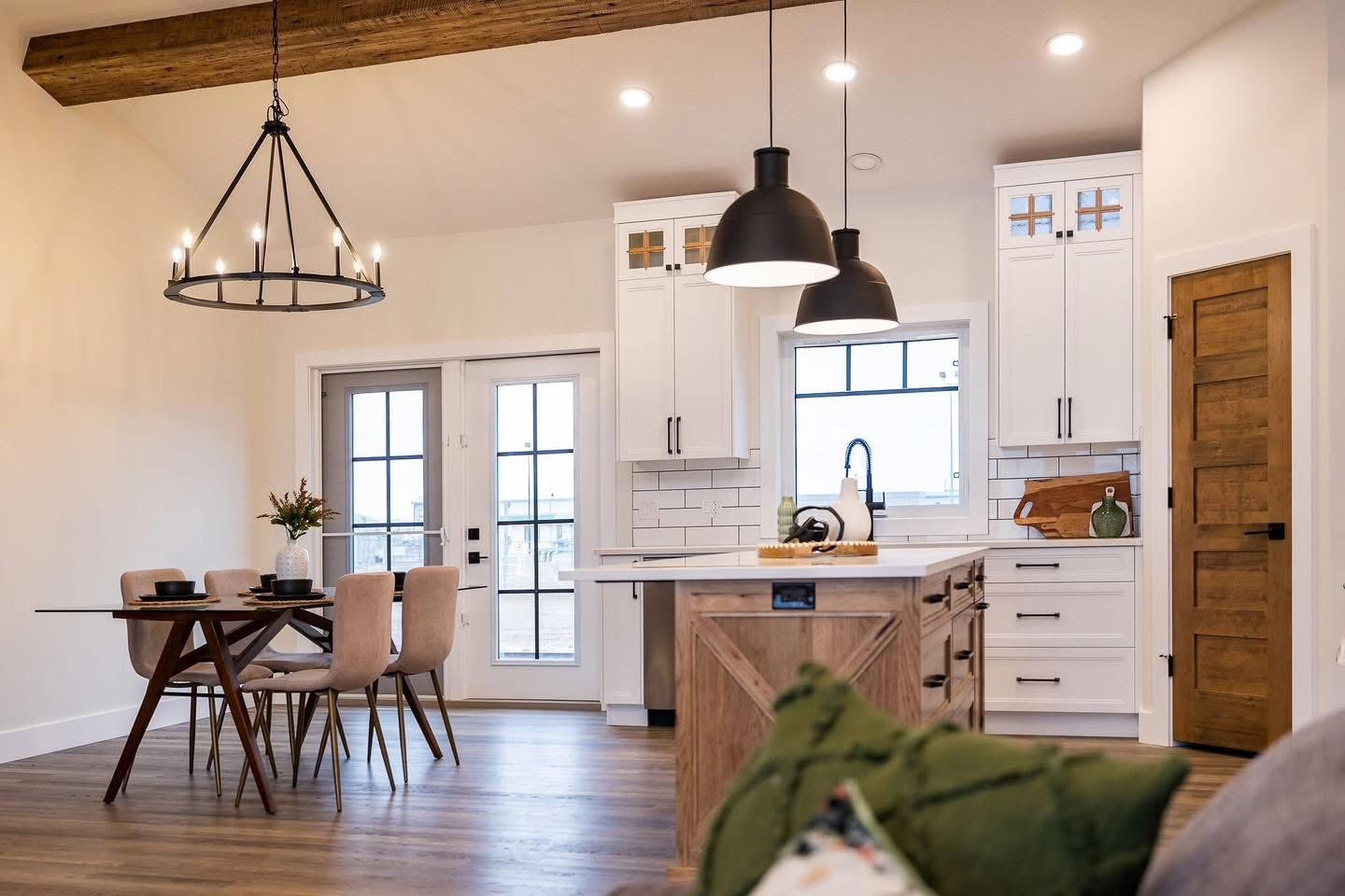 Modern farmhouse kitchen and dining area with wooden beams, chandelier, and pendant lights. Features white cabinets, a central island, and a cozy dining table.