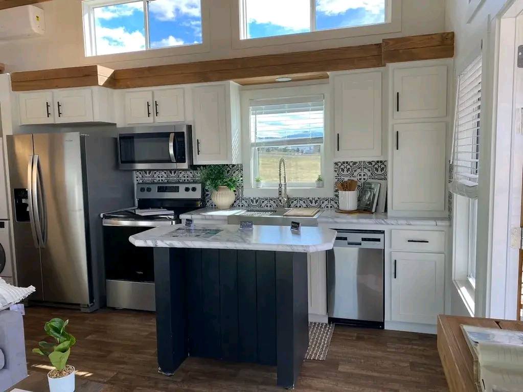 Bright, modern kitchen with white cabinets, stainless steel appliances, a central island, and farmhouse decor. Sunlight streams through a window.