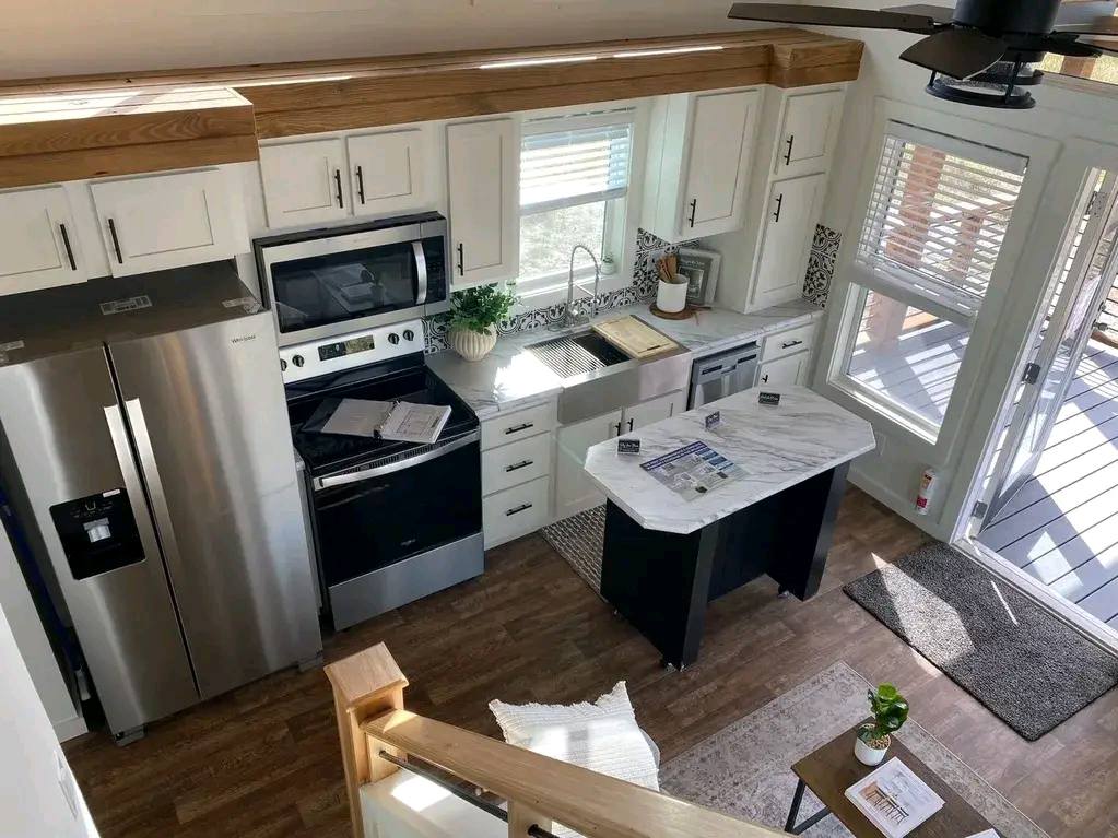 Modern kitchen with white cabinets, stainless steel appliances, and a center island. Sunlight streams through large windows, creating a bright, welcoming atmosphere.