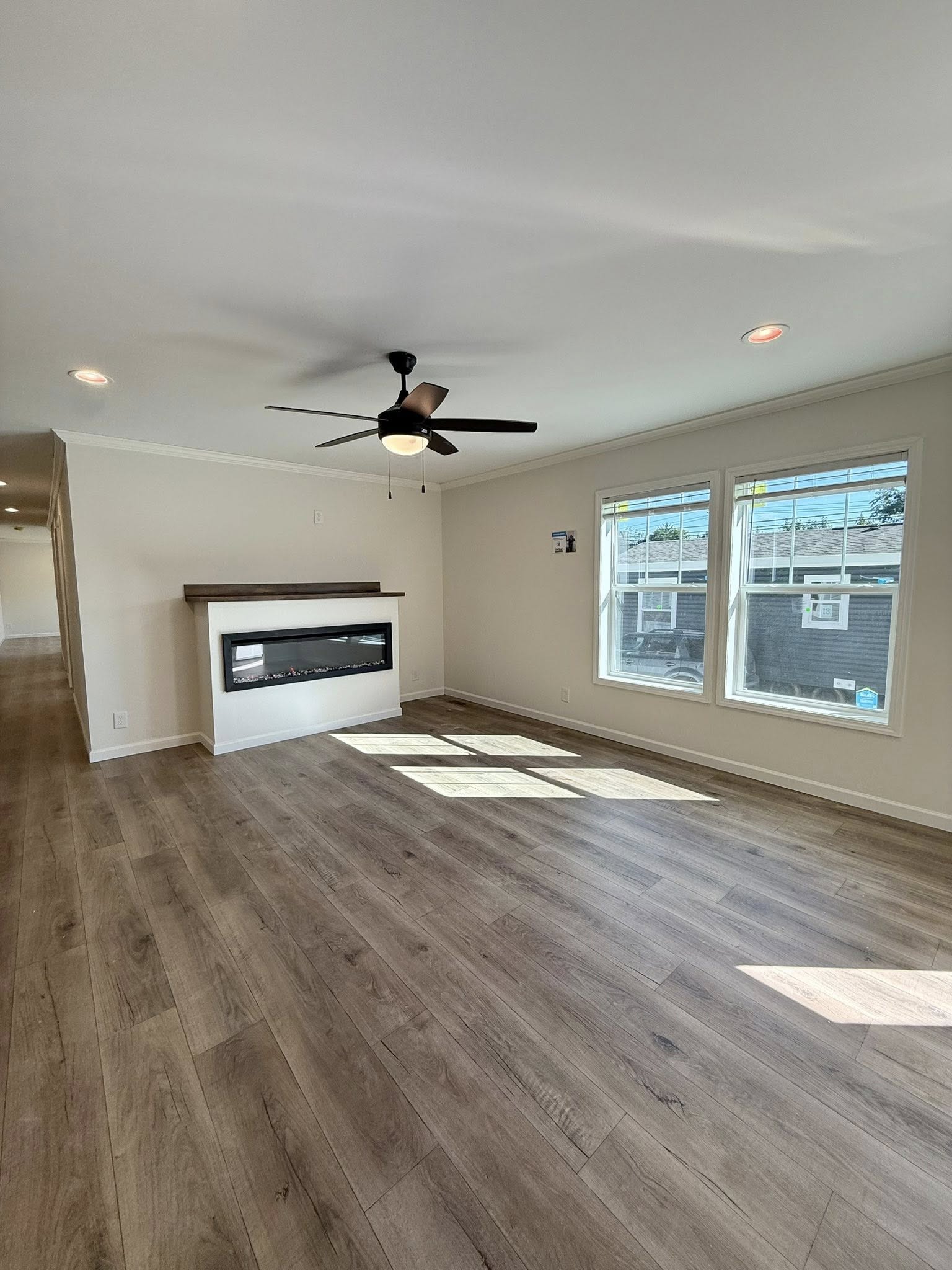 Spacious living room with light wood flooring, modern electric fireplace, ceiling fan, and large windows letting in natural light, creating a bright atmosphere.