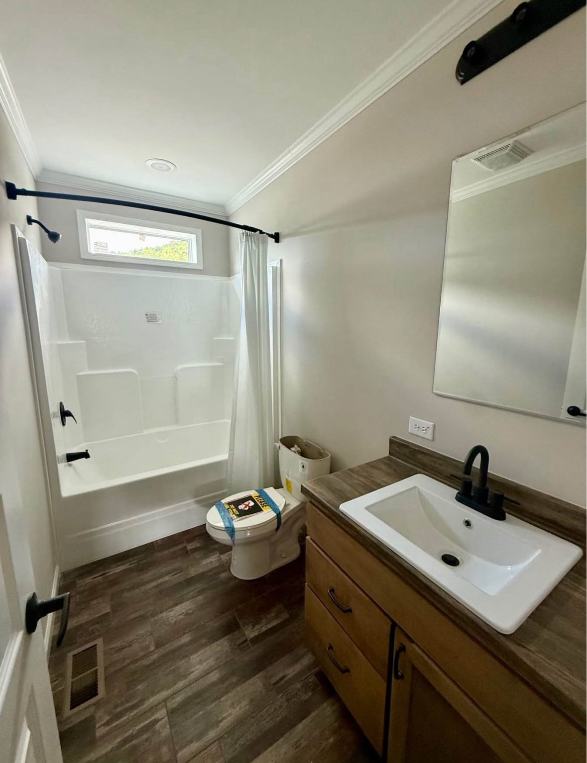 Modern bathroom with wood floors, featuring a white bathtub with a curtain, toilet with cleaning supplies on top, and a wooden vanity with a sleek faucet.