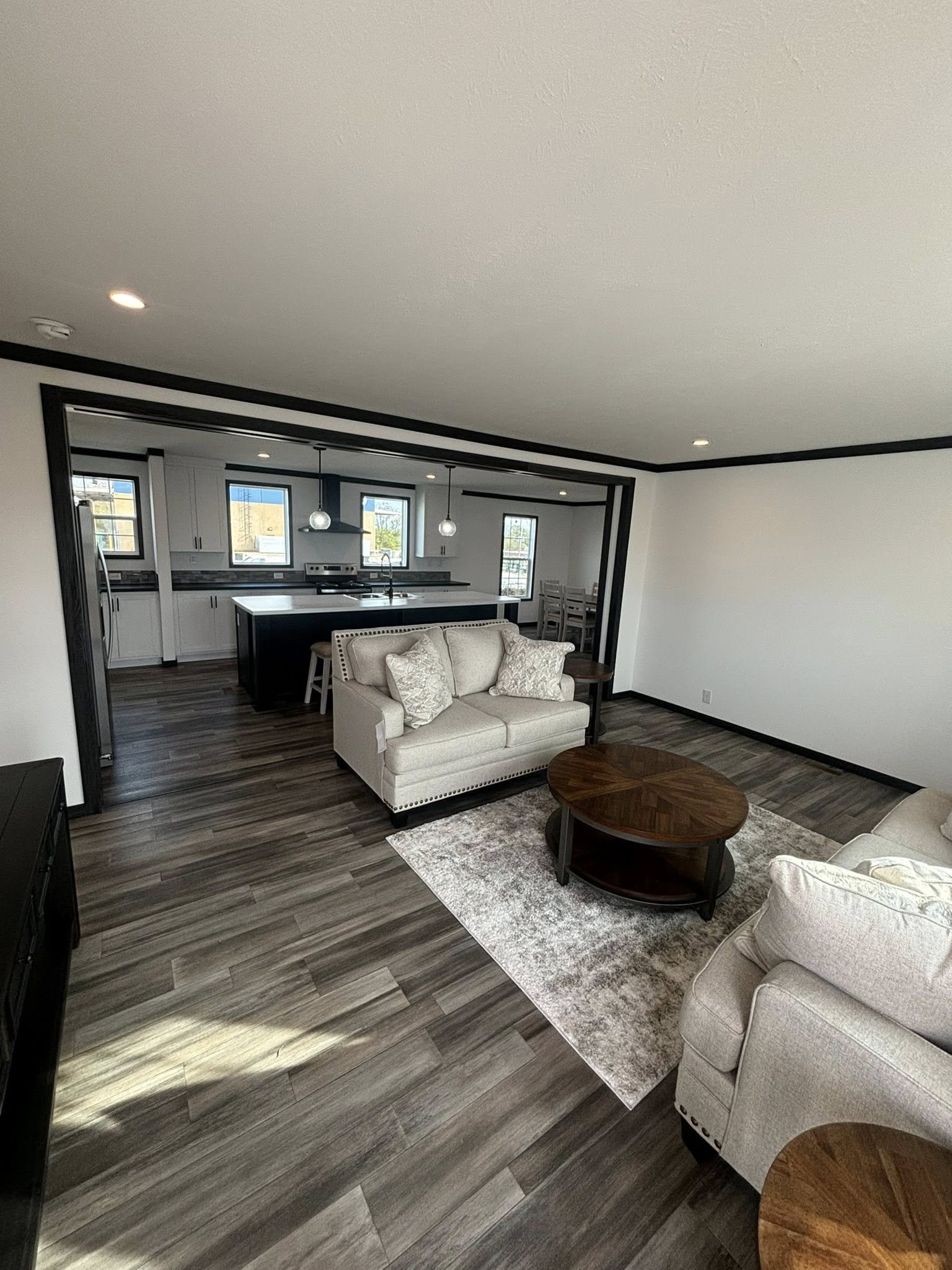 Modern open-concept living space with two beige sofas, a round wooden coffee table on a gray rug, and a kitchen island in the background. Bright and inviting.
