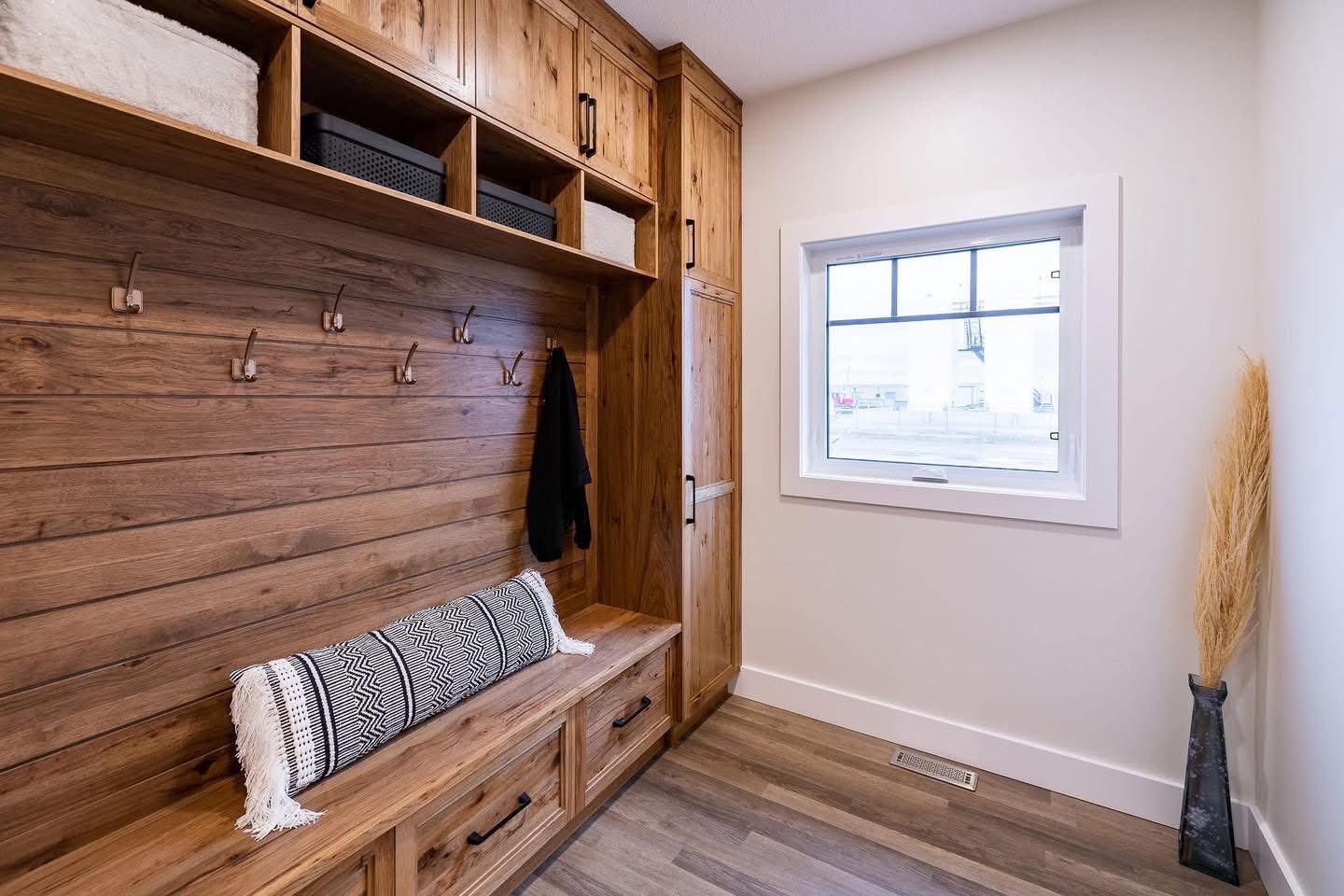 A cozy mudroom with wooden cabinets, a bench, patterned cushion, and wall hooks on the left. A window and tall vase with dried grasses are on the right.
