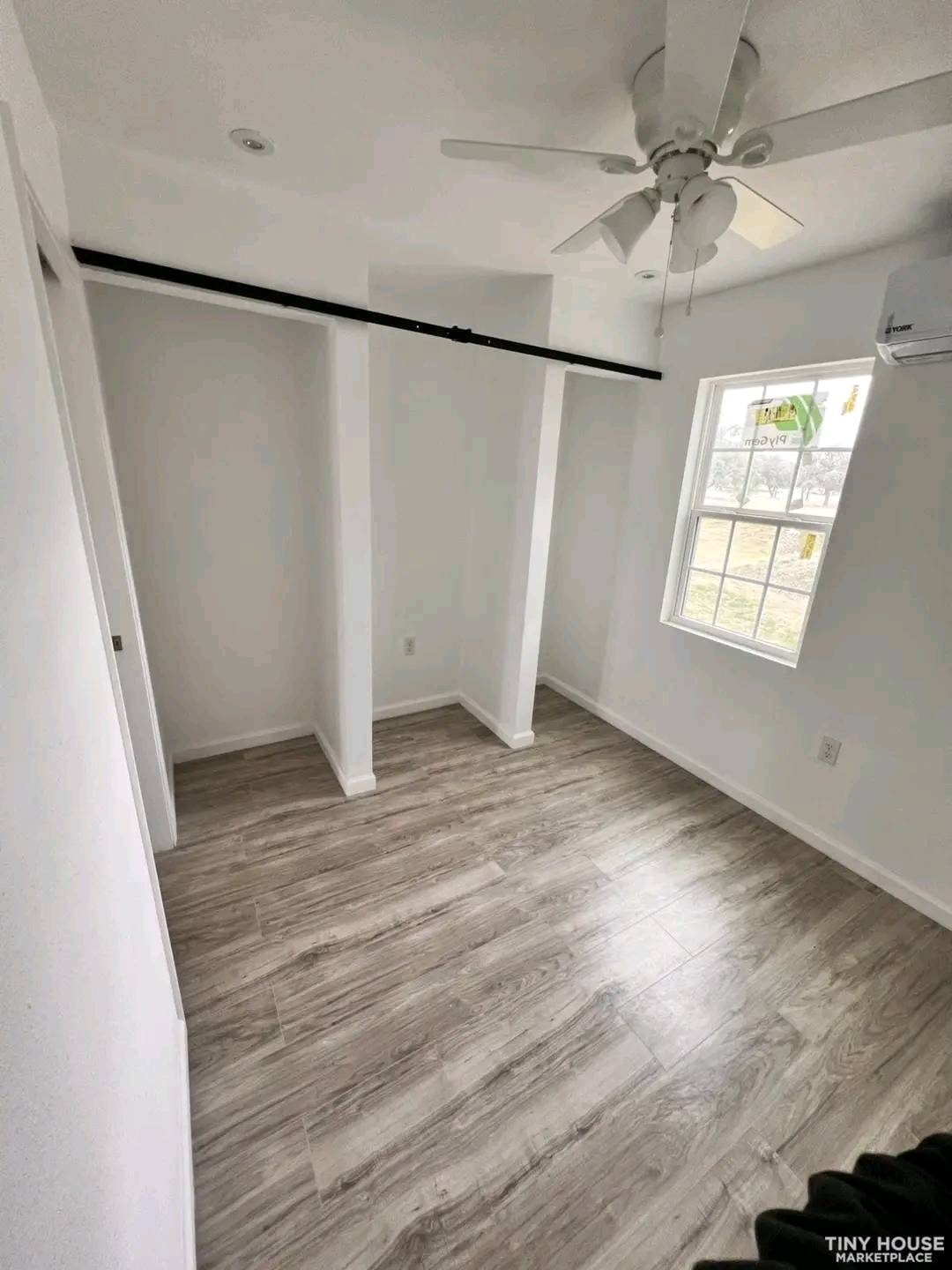 Small, bright room with light wood flooring, white walls, and a ceiling fan. A window on the right allows natural light, creating an airy feel.