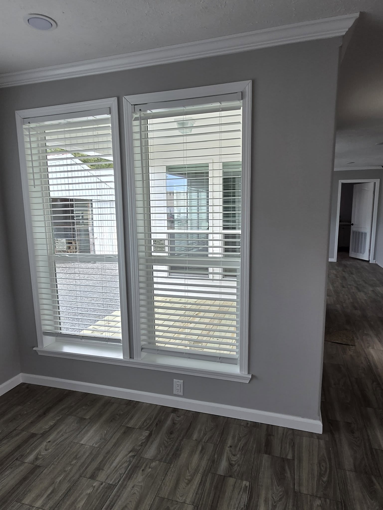 Two large windows with white blinds in a modern room with gray walls and wood flooring. Natural light streams through, creating a calm ambiance.