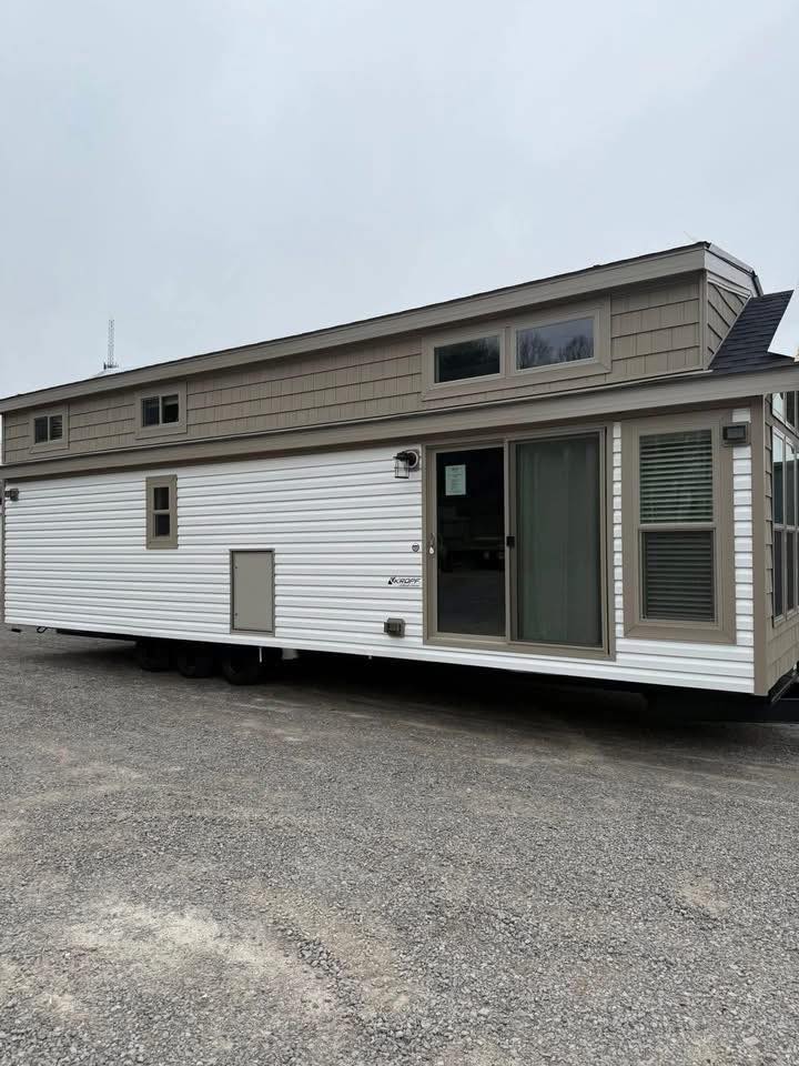Modern tiny home on wheels with white siding and gray trim, featuring multiple windows and a sliding glass door, set against a cloudy sky.